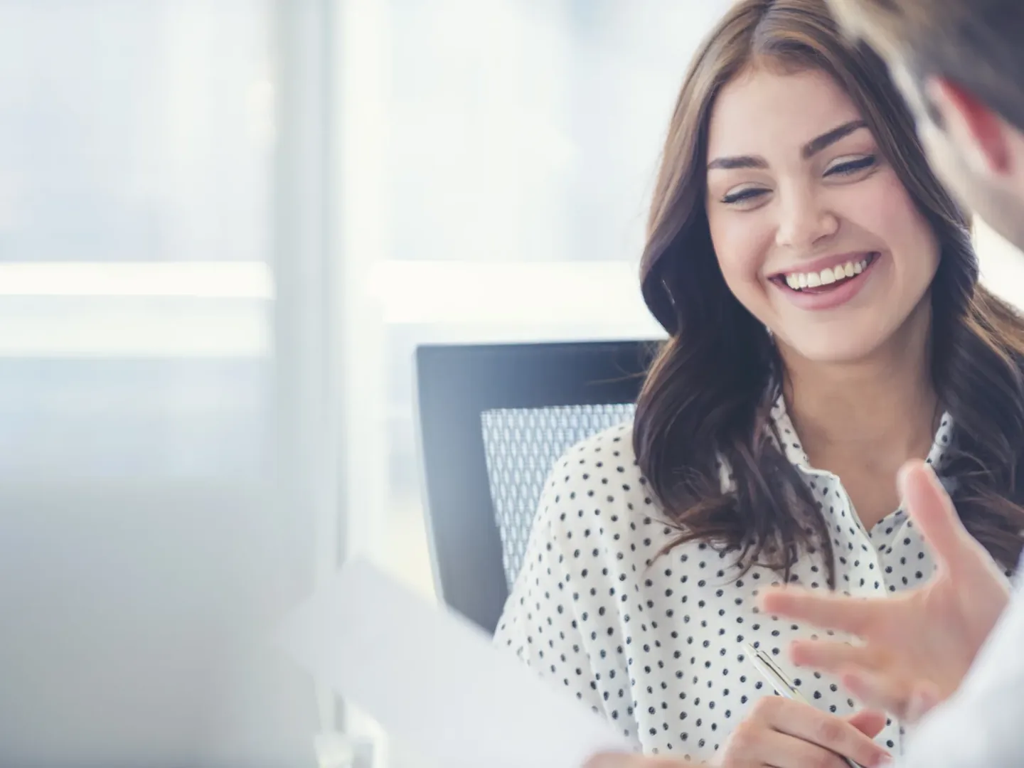 A woman smiling and gesturing mid-conversation across a desk in a bright office