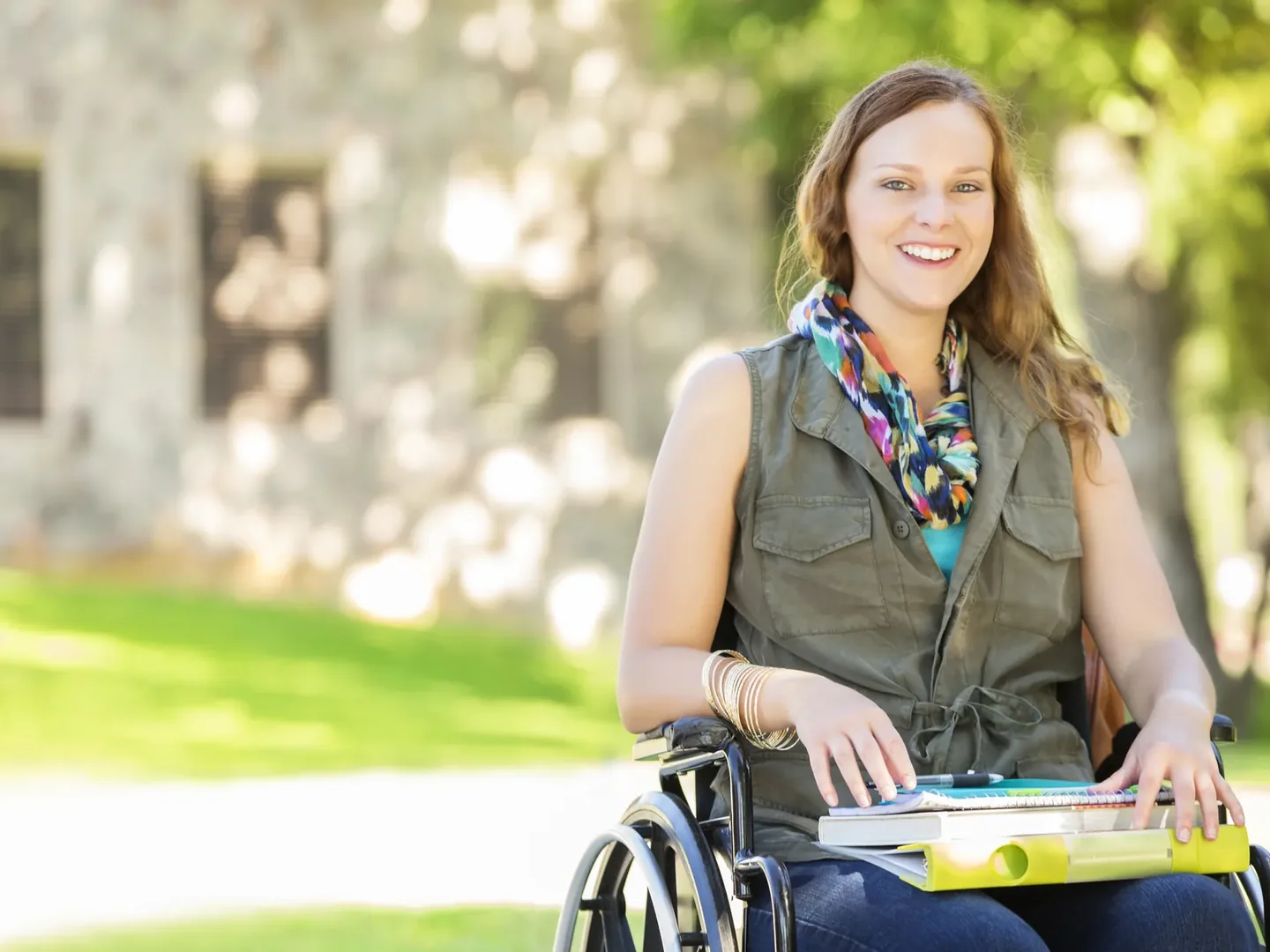 A woman in a wheelchair outdoors on a sunny path, holding folders and smiling