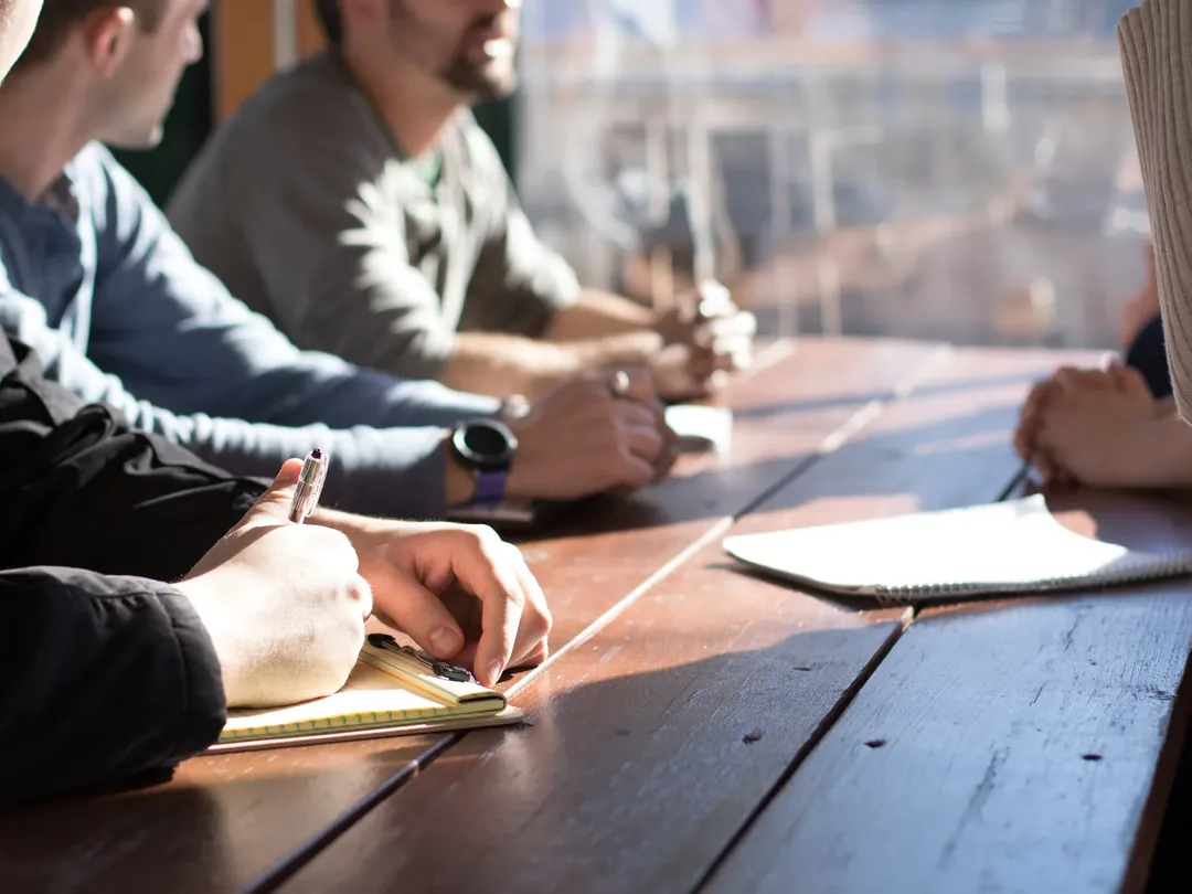 Four people around a wooden table in sunlight, taking notes in notebooks during a meeting.