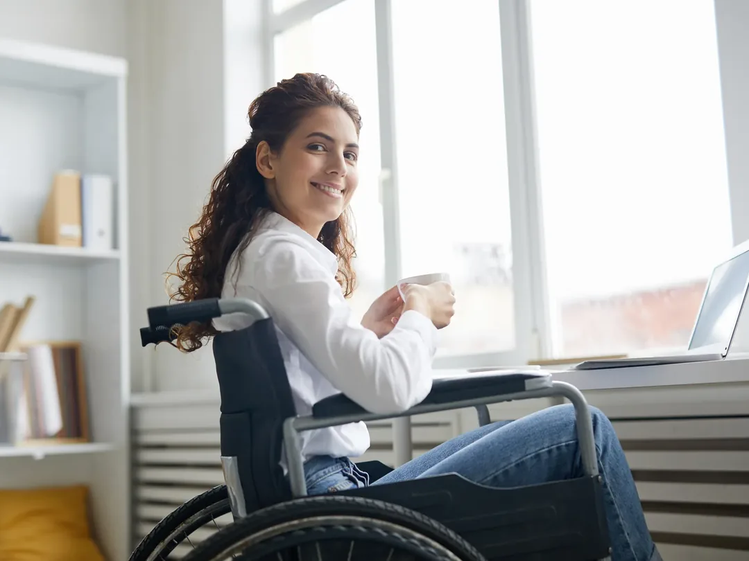 A woman in a wheelchair at a desk by a window, holding a mug, with a laptop open beside her.