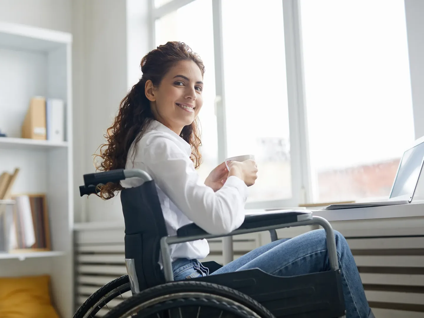 A woman in a wheelchair at a desk by a window, holding a mug, with a laptop open beside her