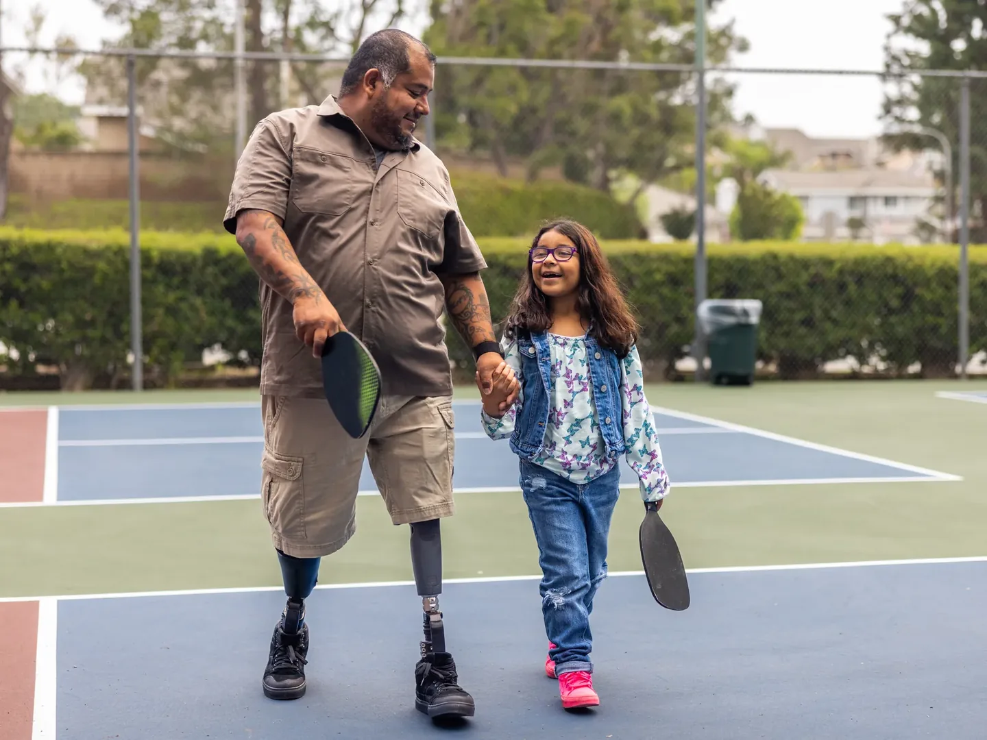 A father and his daughter walking hand in hand off a tennis court, holding bats and smiling
