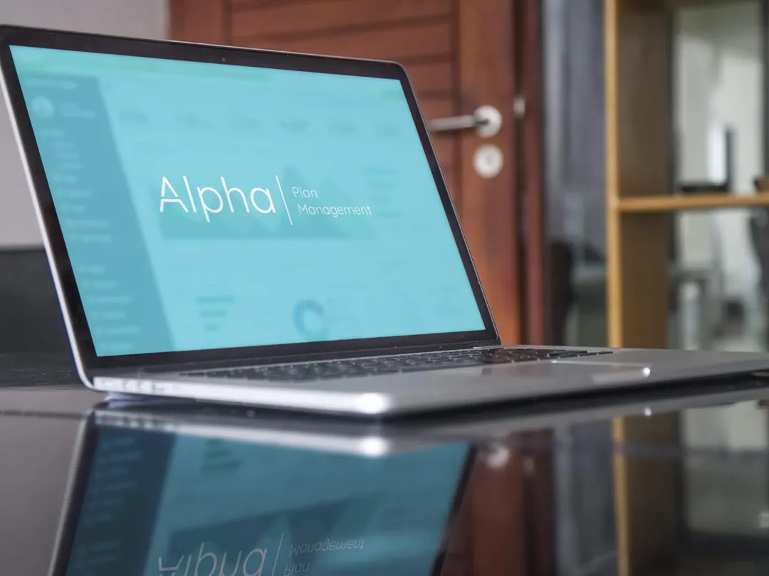 A laptop on a dark desk showing the Alpha Plan Management dashboard.